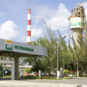 Entrada de uma unidade da Petrobras com uma guarita com o logotipo da empresa e, ao fundo, duas chamin&eacute;s industriais e uma torre com o selo "BR"