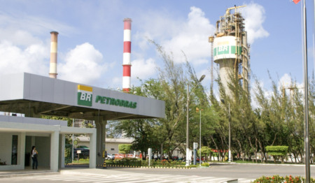 Entrada de uma unidade da Petrobras com uma guarita com o logotipo da empresa e, ao fundo, duas chaminés industriais e uma torre com o selo "BR"