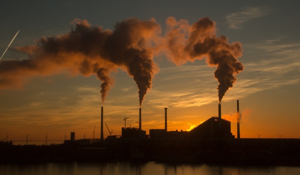 Silhueta de um complexo industrial emitindo fumaça de suas chaminés contra um céu de pôr do sol alaranjado. Silhueta de um complexo industrial emitindo fumaça de suas chaminés contra um céu de pôr do sol alaranjado.