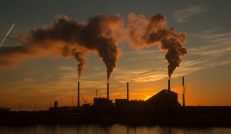 Silhueta de um complexo industrial emitindo fumaça de suas chaminés contra um céu de pôr do sol alaranjado. Silhueta de um complexo industrial emitindo fumaça de suas chaminés contra um céu de pôr do sol alaranjado.