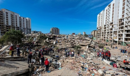Foto de área com escombros causados pelo terremoto, cercada por alguns prédios altos também danificados. Equipes de resgate e outras pessoas estão sobre os escombros
