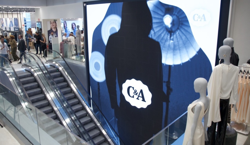 Interior de loja da C&A, mostra escadas rolantes, manequins e pessoas, com um telão com am silhueta de uma mulher e o logo da loja Interior de loja da C&A, mostra escadas rolantes, manequins e pessoas, com um telão com am silhueta de uma mulher e o logo da loja