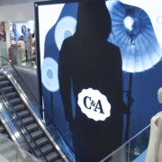 Interior de loja da C&A, mostra escadas rolantes, manequins e pessoas, com um tel&atilde;o com am silhueta de uma mulher e o logo da loja
