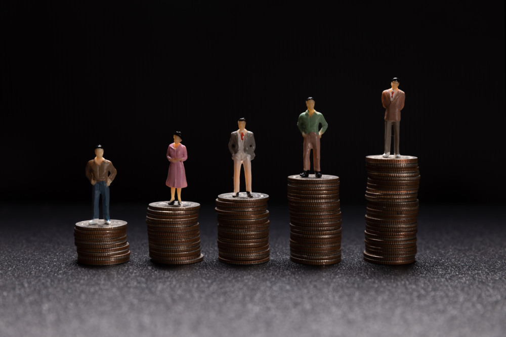 A imagem mostra cinco bonecos em miniatura, representando pessoas de negócios, em pé sobre pilhas de moedas de diferentes alturas, formando uma composição em degraus crescentes da esquerda para a direita A imagem mostra cinco bonecos em miniatura, representando pessoas de negócios, em pé sobre pilhas de moedas de diferentes alturas, formando uma composição em degraus crescentes da esquerda para a direita