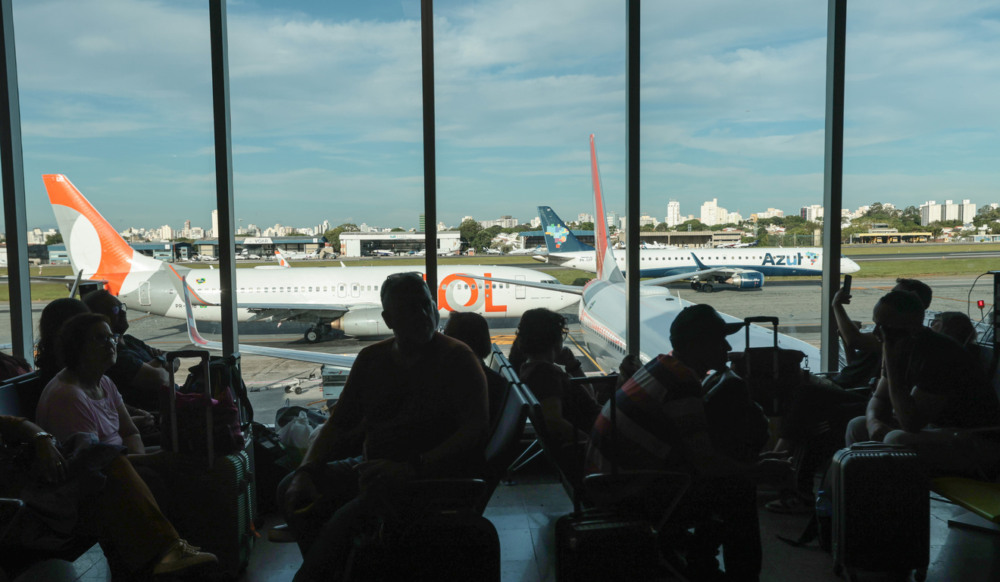 Passageiros na área de espera do terminal de aeroporto. Atrás, a vista de duas aeronaves comerciais, uma da Gol e uma da Azul Passageiros na área de espera do terminal de aeroporto. Atrás, a vista de duas aeronaves comerciais, uma da Gol e uma da Azul