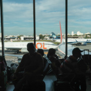 Passageiros na &aacute;rea de espera do terminal de aeroporto. Atr&aacute;s, a vista de duas aeronaves comerciais, uma da Gol e uma da Azul