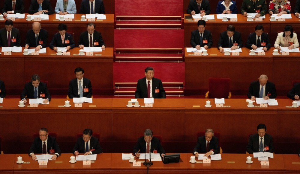 Xi Jinping senta numa mesa no meio da foto, cercado por outros membros do parlamento chinês sentados em mesas. Todos escutam um discurso Xi Jinping senta numa mesa no meio da foto, cercado por outros membros do parlamento chinês sentados em mesas. Todos escutam um discurso