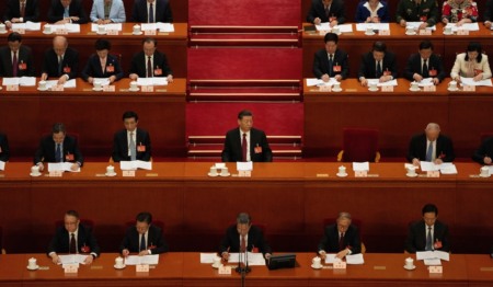 Xi Jinping senta numa mesa no meio da foto, cercado por outros membros do parlamento chinês sentados em mesas. Todos escutam um discurso Xi Jinping senta numa mesa no meio da foto, cercado por outros membros do parlamento chinês sentados em mesas. Todos escutam um discurso