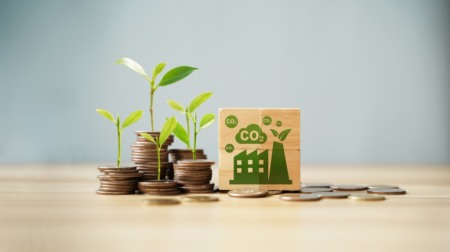 Pilhas de moedas com folhas de plantas saindo delas ao lado de um bloco com uma imagem de fábrica e nuvens de CO2 Pilhas de moedas com folhas de plantas saindo delas ao lado de um bloco com uma imagem de fábrica e nuvens de CO2