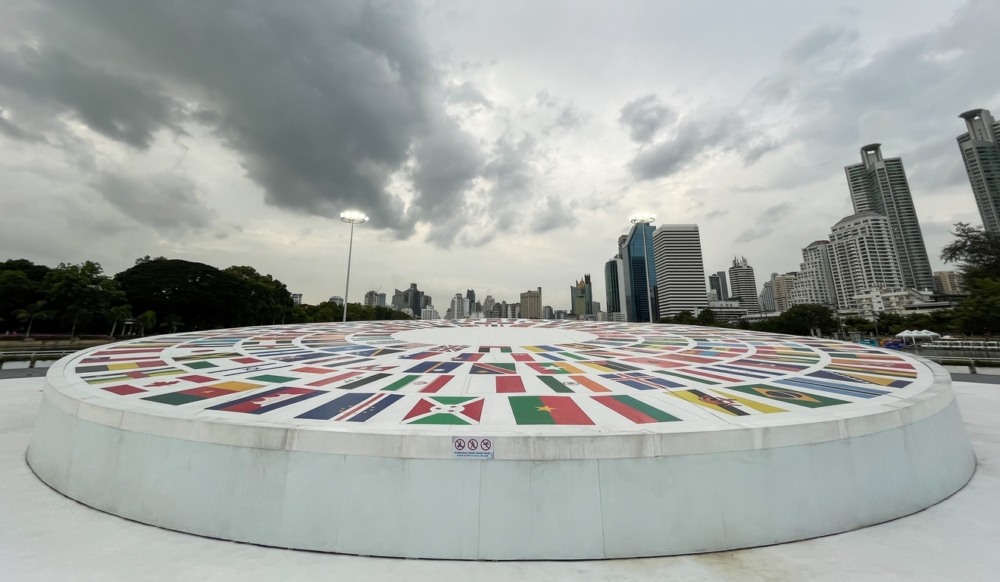 Monumento circular em praça com imagens de bandeiras de vários países. Ao fundo estão prédios, e o véu está carregado de nuvens Monumento circular em praça com imagens de bandeiras de vários países. Ao fundo estão prédios, e o véu está carregado de nuvens
