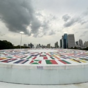Monumento circular em pra&ccedil;a com imagens de bandeiras de v&aacute;rios pa&iacute;ses. Ao fundo est&atilde;o pr&eacute;dios, e o v&eacute;u est&aacute; carregado de nuvens