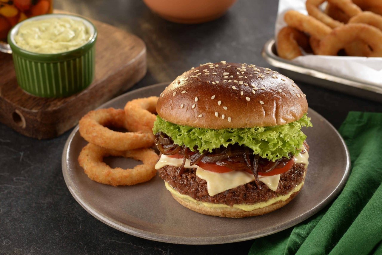 Cheeseburguer com hamburguer plant-based, queijo, salada de alface e tomate, ao lado de anéis de cebola, sobre um prato