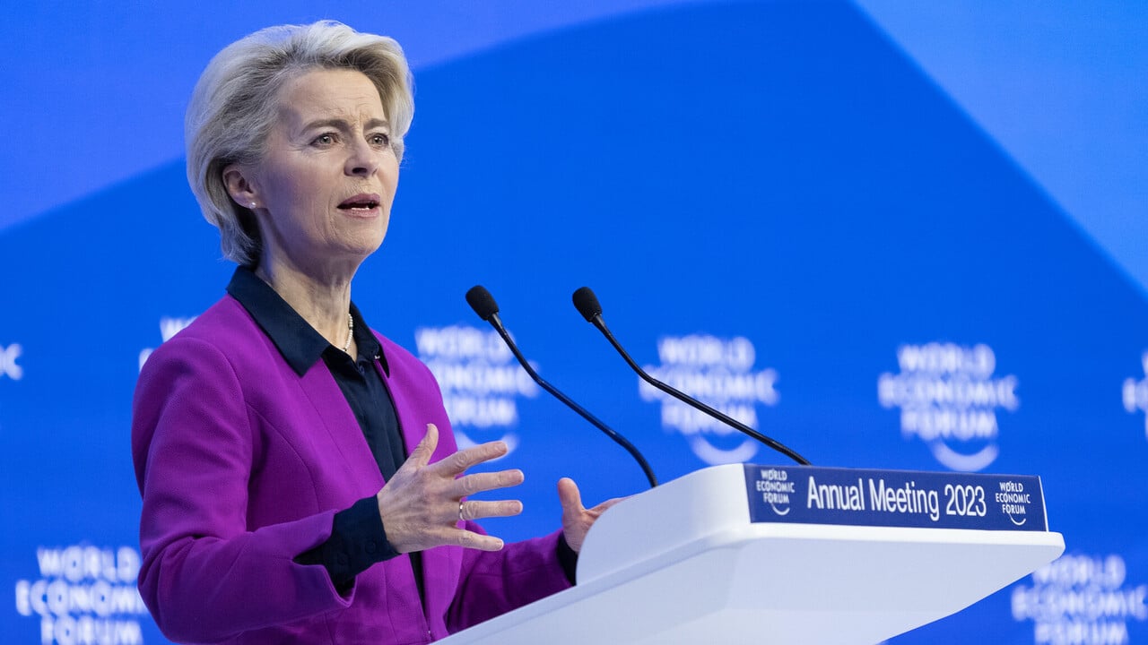 Mulher branca, com cabelos curtos e loiros claros, usando terno púrpura com blusa preta embaixo. Ela fala em palanque, com fundo azul atrás do Fórum Econômico Mundial.