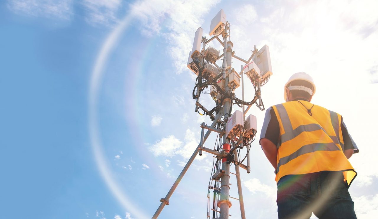 Helmeted asian male engineer works in the field with a telecommunication tower that controls cellular electrical installations to inspect and maintain 5G networks installed on high-rise buildings.